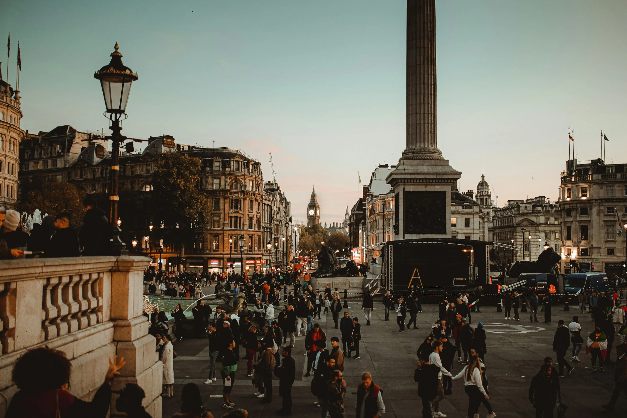 Crowd gathered in Trafalgar Square in London during dusk, with Big Ben clock tower visible in the background and a prominent large column monument in the center of the square.