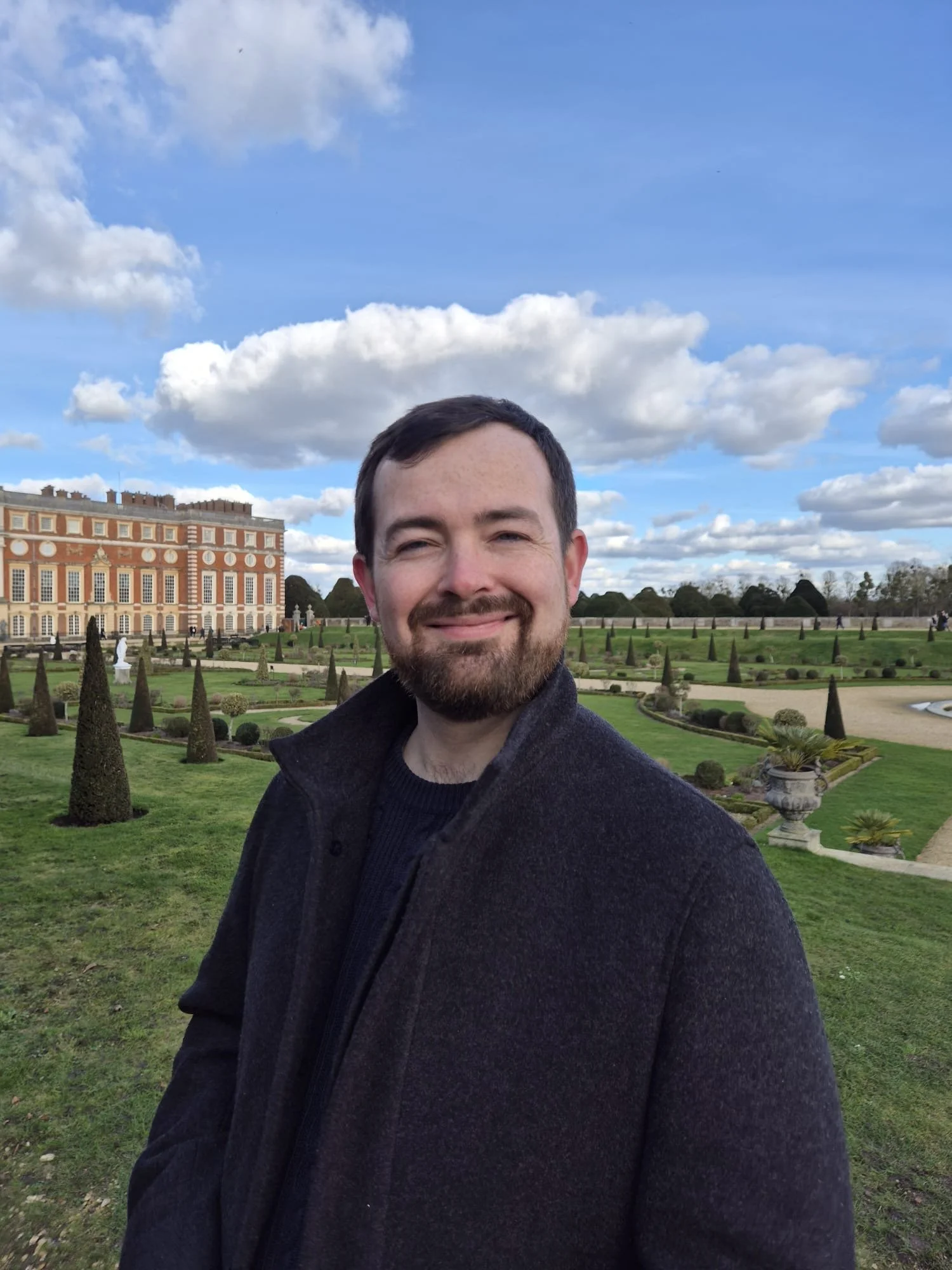 A man smiling outdoors in front of a garden with trimmed bushes and statues, with a large historic building and a partly cloudy sky in the background.