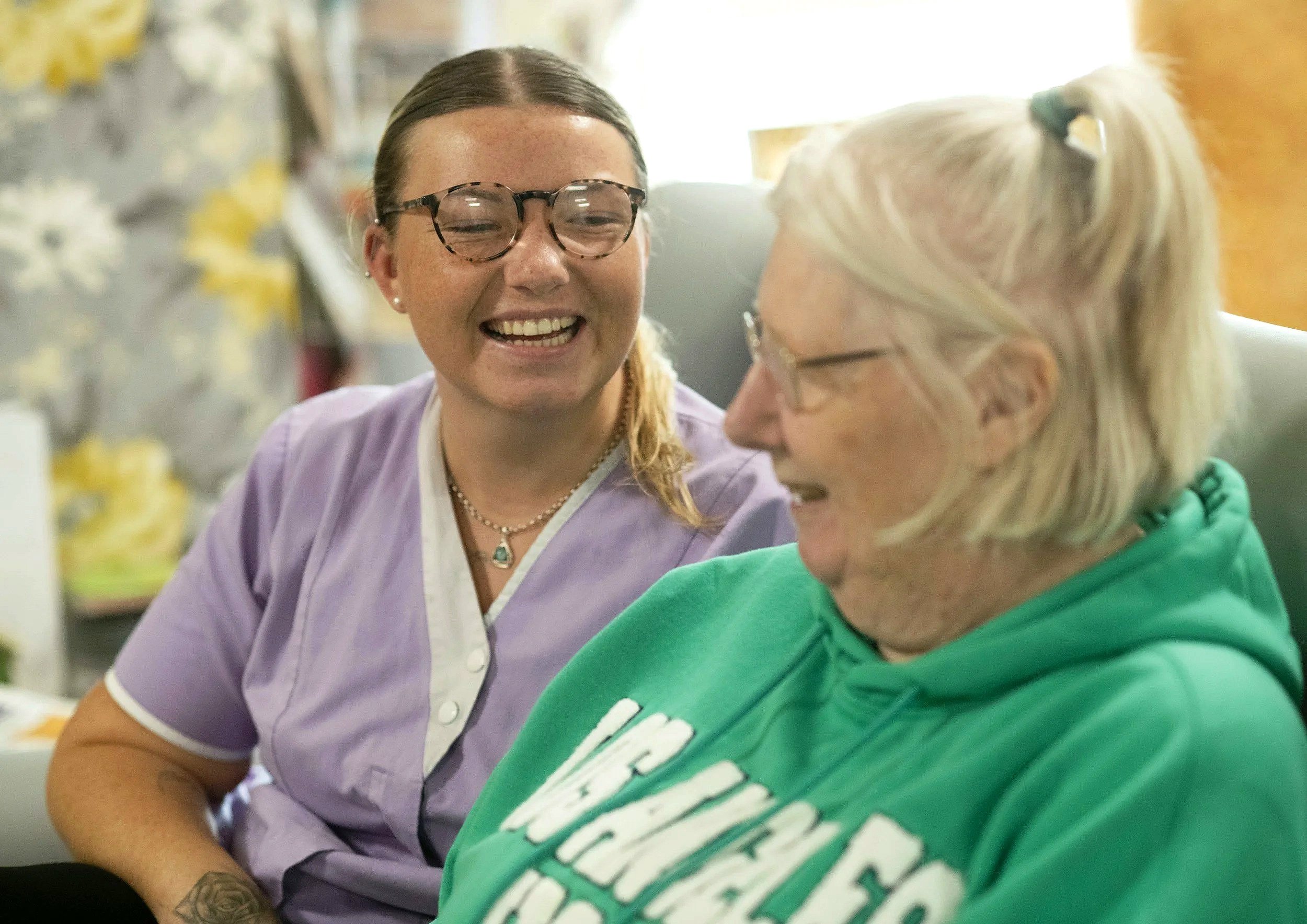 Two women are sitting close to each other, smiling and sharing a joyful moment. One woman has brown hair, glasses, and is wearing a purple shirt, while the other has blond hair tied in a ponytail and is wearing a green hoodie.