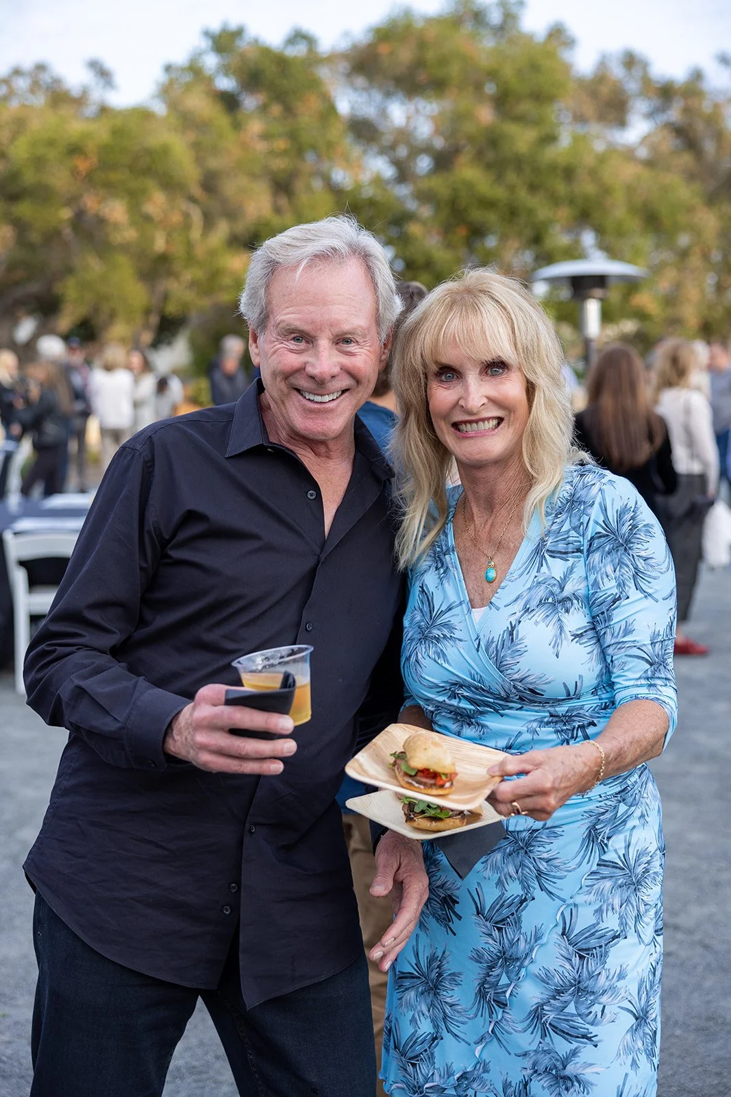 A smiling older man in a black shirt holding a drink, standing next to a smiling older woman in a blue floral dress holding two mini sandwiches at an outdoor event, with trees and other people in the background.