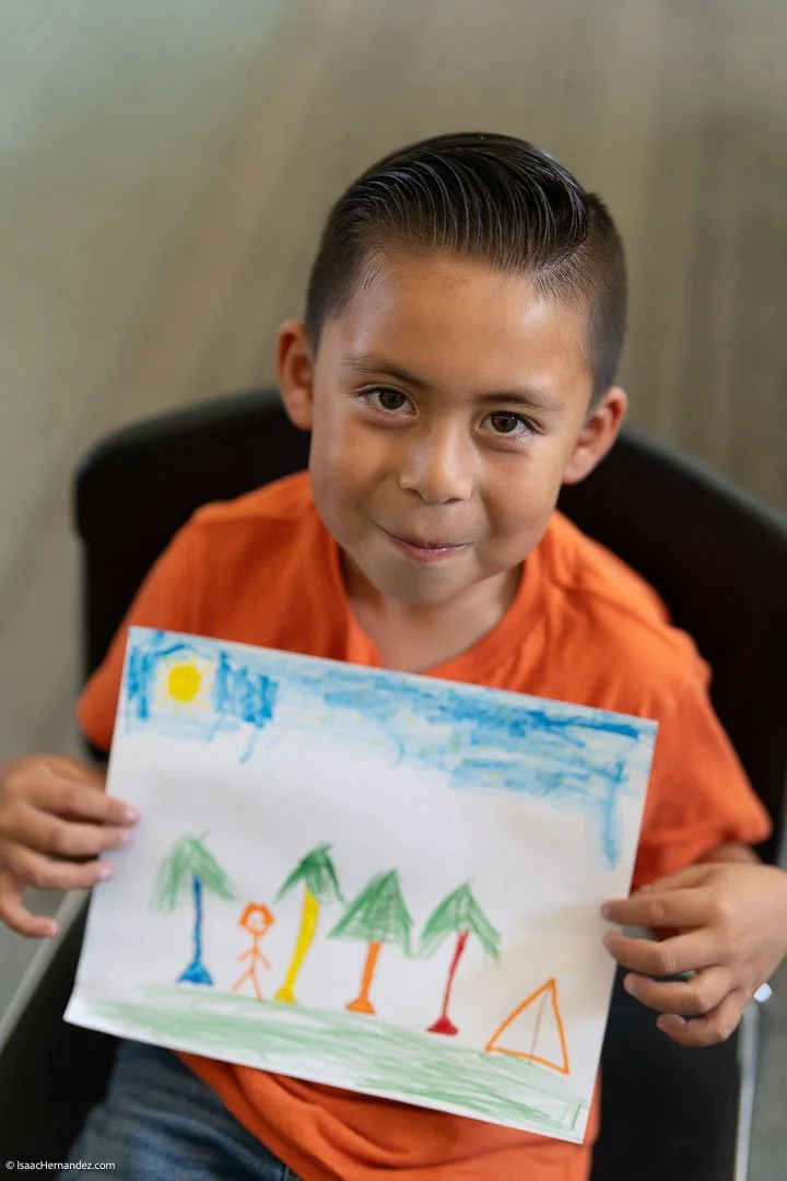 A young boy in an orange shirt sitting down and holding a colorful drawing of a sunny day with trees, a person, and a tent.