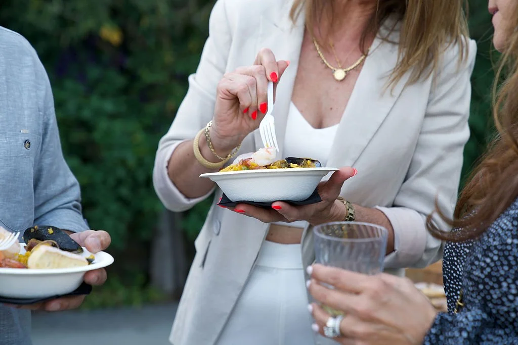 People at an outdoor gathering holding plates of food and a glass, engaging in conversation.