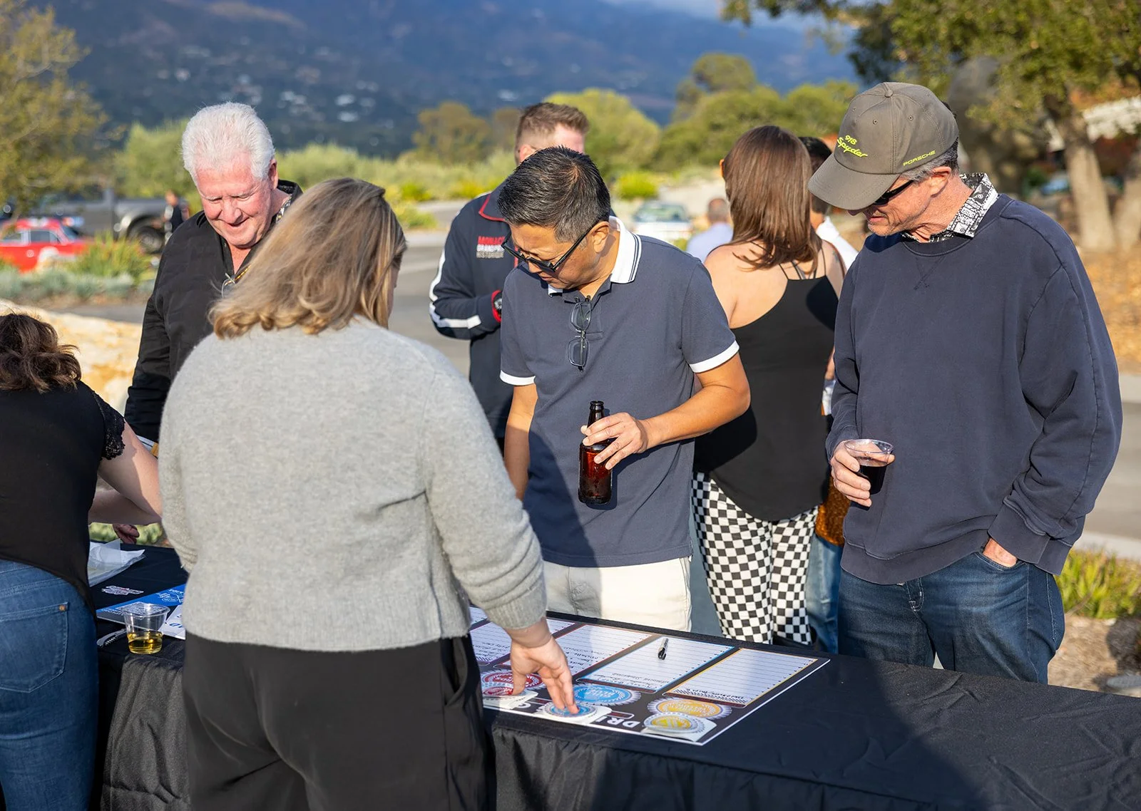 Group of people gathered around an outdoor table, looking at papers and conversing, during a sunny day with trees and parked cars in the background.