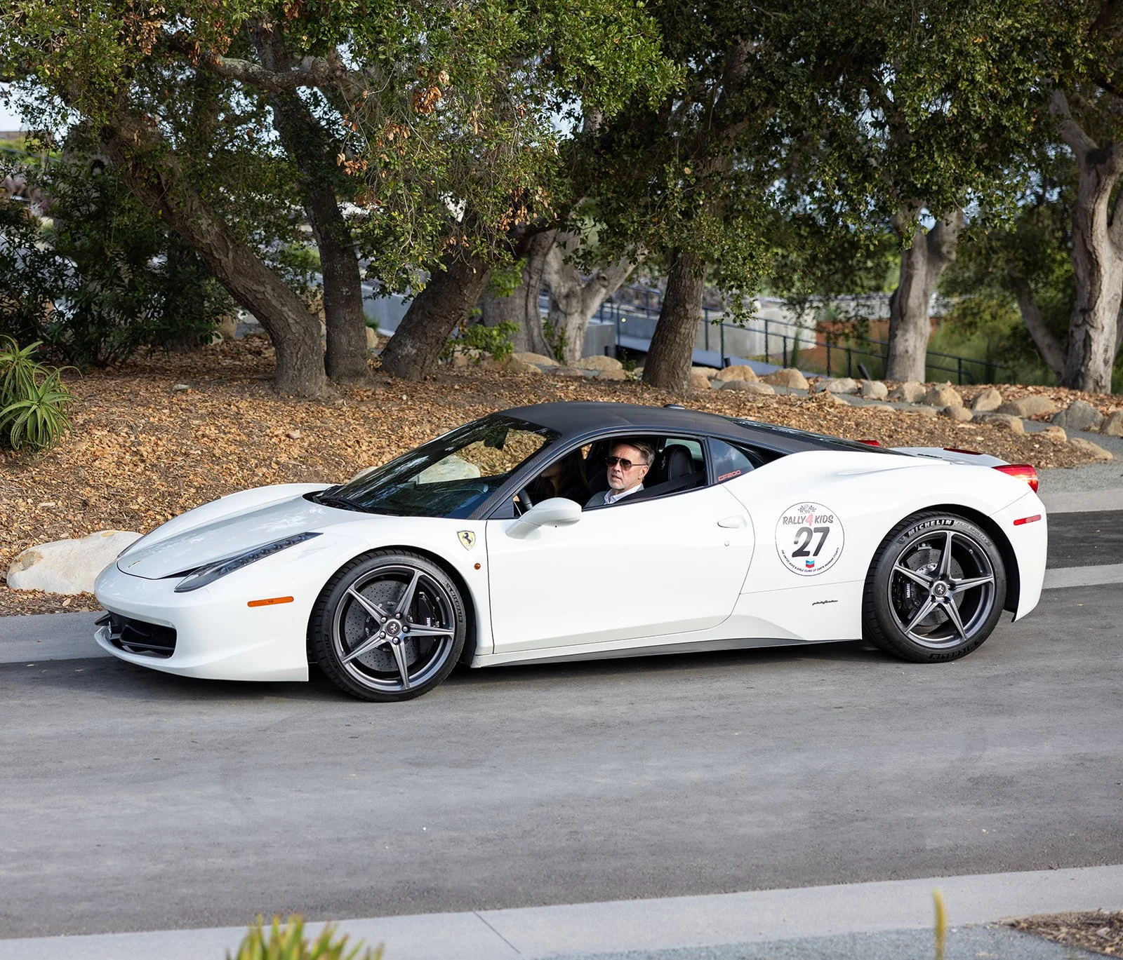 A white Ferrari sports car parked on the side of a street with a man inside wearing sunglasses and a suit. The car has racing decals on the side, including the number 27 and the logo for Rally Kids.