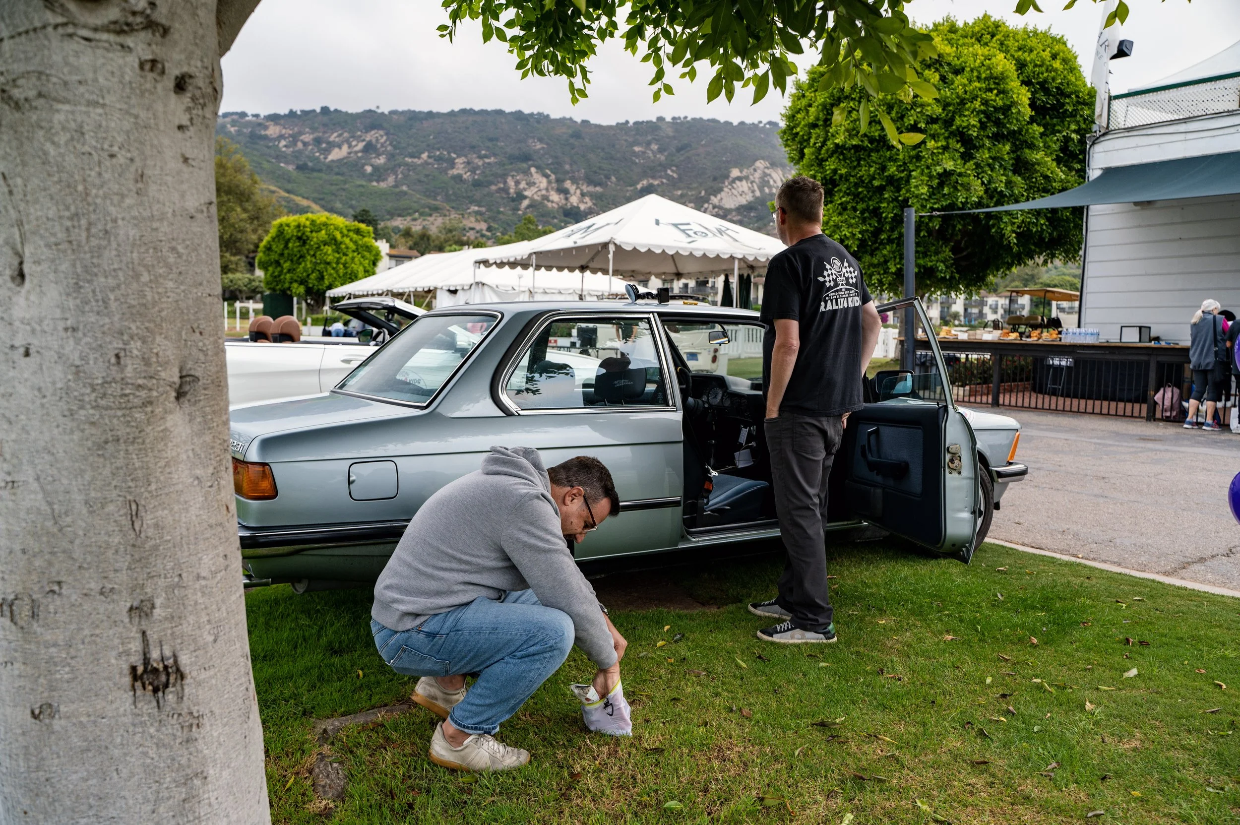 Man in glasses crouching near grass and a man in a black shirt standing near an open car door at an outdoor event with tents and trees.