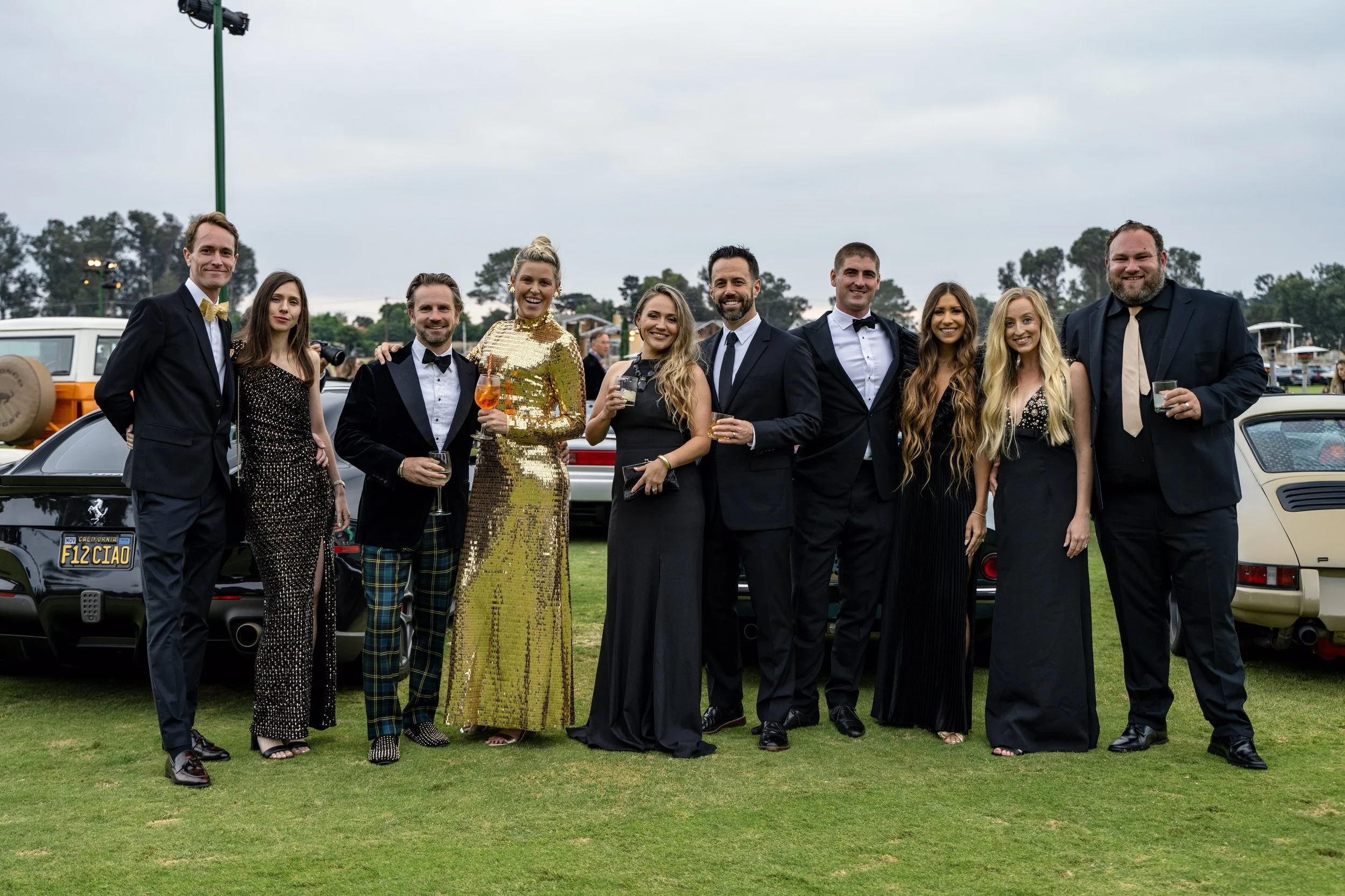 A group of ten people dressed in formal attire standing together outdoors at a car show or event, holding drinks and smiling.