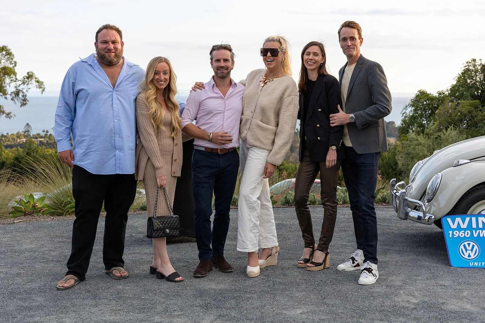 A group of seven people standing outdoors on a gravel area, smiling at the camera with scenic greenery and ocean in the background, next to a vintage white Volkswagen Beetle with a sign that says 'WIN 1960 VW'.