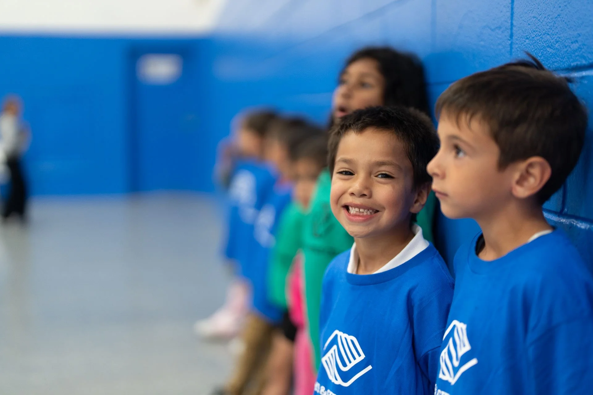 Smiling young boy in a blue shirt standing in line with other children against a blue wall in an indoor setting.