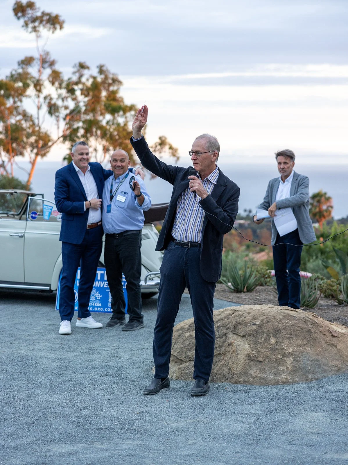 A man in a suit speaking into a microphone at an outdoor event, with three men in the background near a vintage white convertible car, outdoors during late afternoon or early evening.