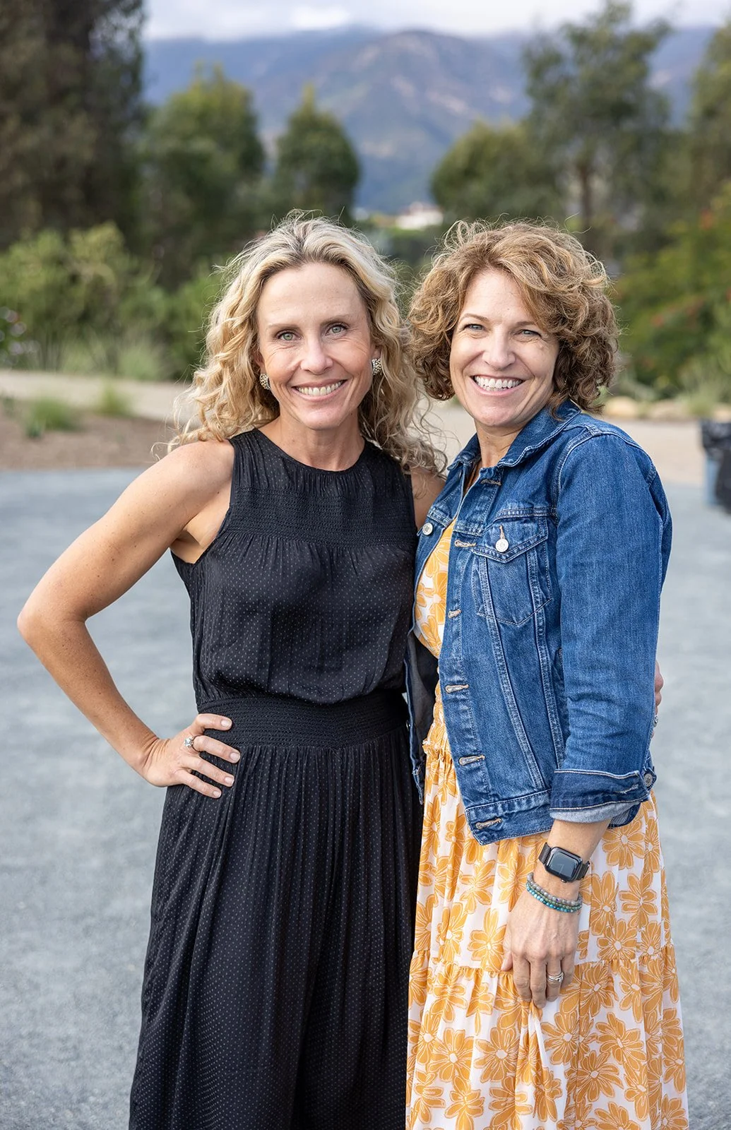 Two women standing outdoors, smiling at the camera. One has curly blonde hair and is wearing a black sleeveless dress; the other has curly brown hair and is wearing a yellow floral dress with a denim jacket.