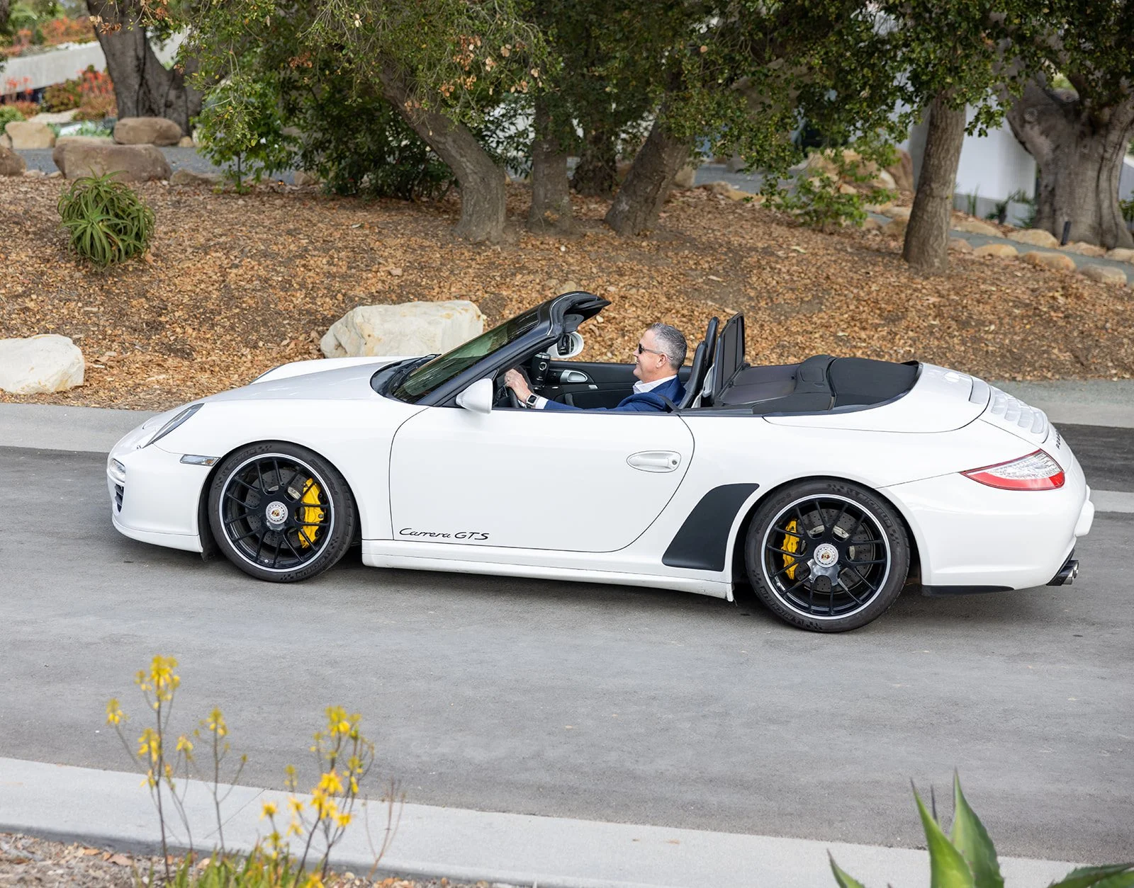 A man driving a white Porsche Carrera GTS convertible on a paved road with a landscaped background including trees and rocks.
