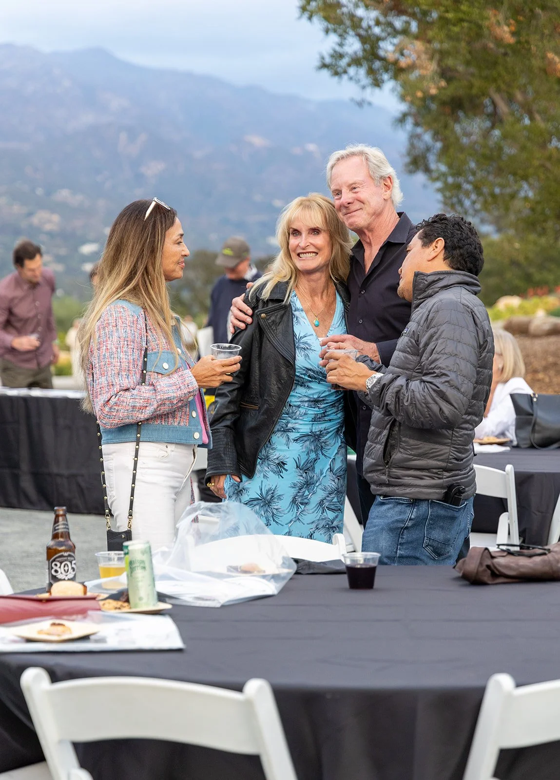 A group of four people, two women and two men, enjoying a social gathering outdoors, with mountains in the background and tables with food and drinks in the foreground.