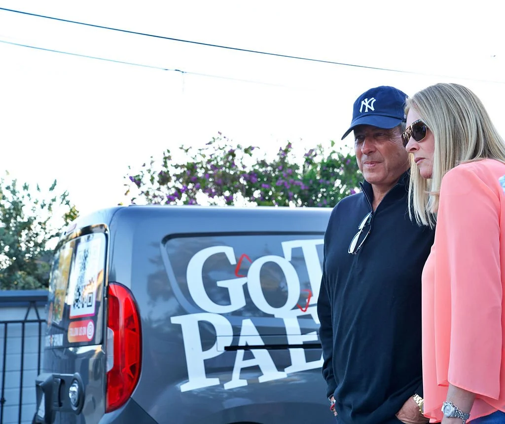 Two casually dressed adults, a man wearing a navy baseball cap and a dark jacket and a woman with blonde hair wearing sunglasses and a peach blazer, standing outdoors near a parked vehicle with a large sign that reads 'Go to Pay'.