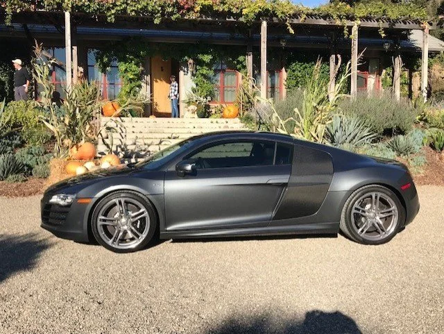 A black Audi R8 sports car parked on a gravel lot with a building, pumpkins, and plants in the background.