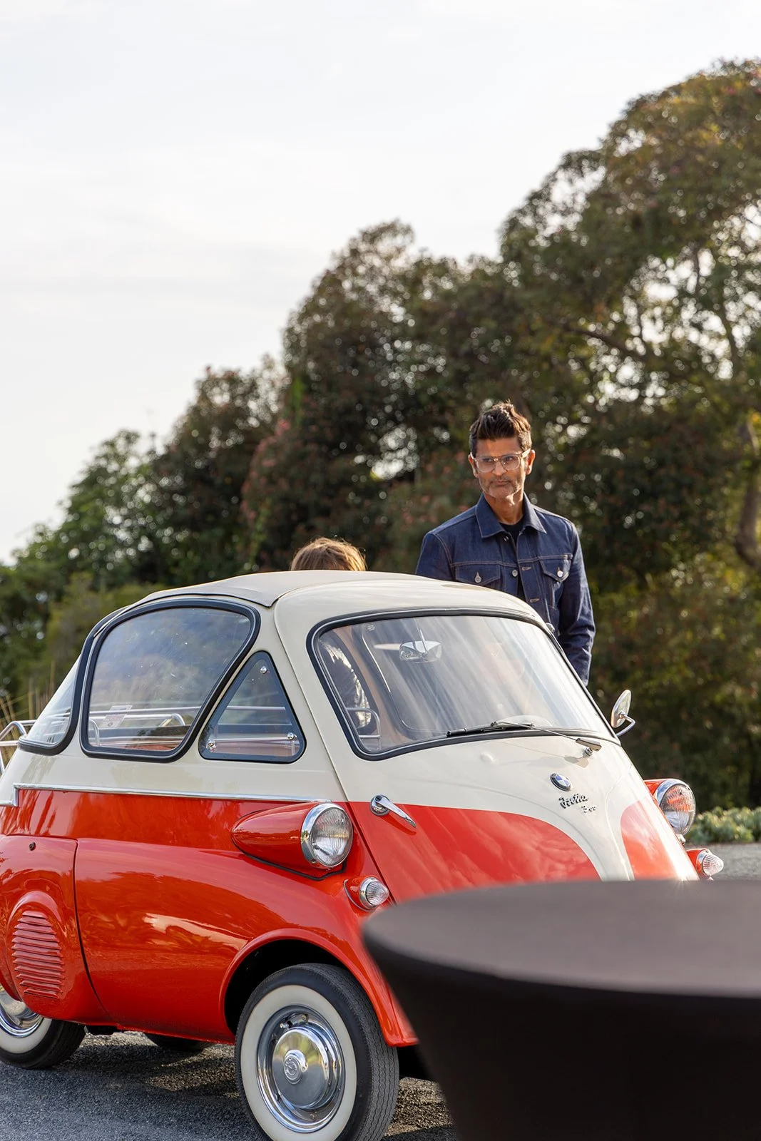 A man with glasses in a denim jacket standing next to a red and white vintage microcar outdoors with trees in the background.