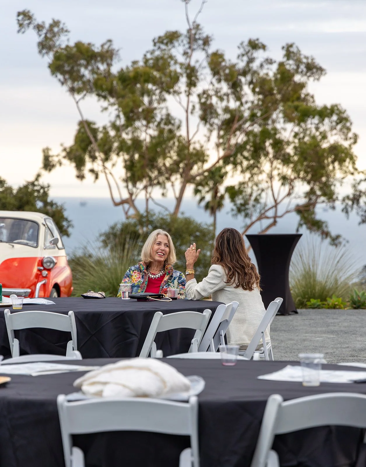 Two women sitting at a black-covered table outdoors, talking and smiling; one woman has blonde hair and wears a colorful blazer, the other has brown hair and wears a white blazer; a vintage van is parked in the background with trees and shrubs.