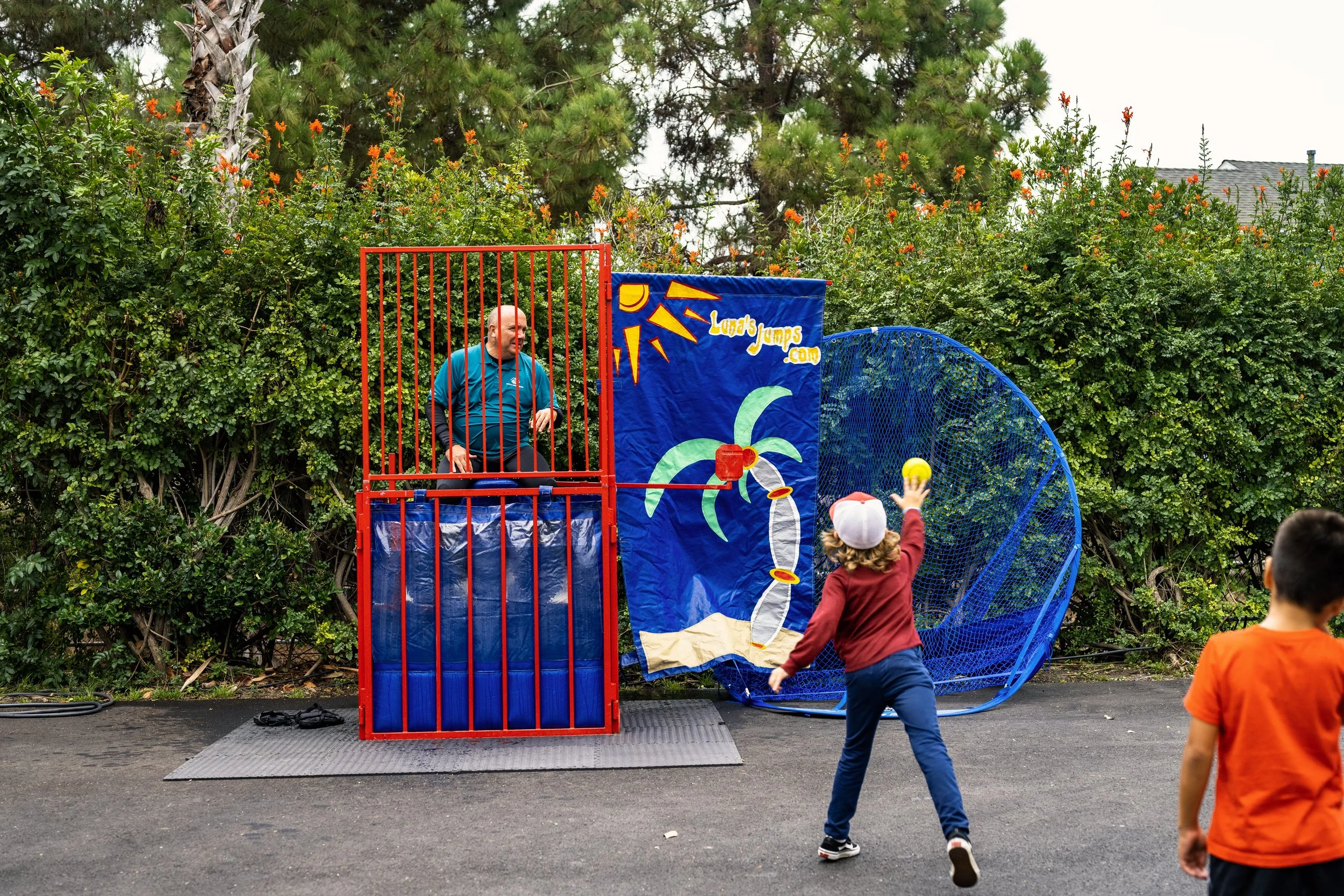 A man stands inside a red cage at a carnival game, aiming a ball at the target. Two children are playing, one jumping with a ball in hand aiming at the target, and a second child watching. The background has bushes and trees, with a blue netted baske