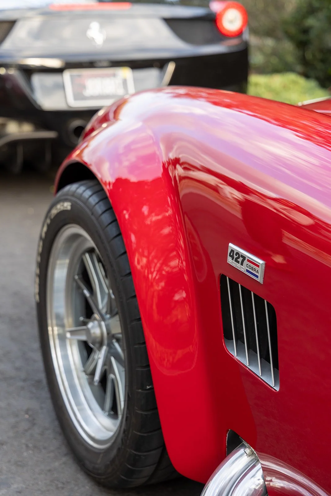 Close-up of a red vintage Chevrolet Corvette with a 427 badge, showing a wheel and side vent, parked outdoors with another car in the background.