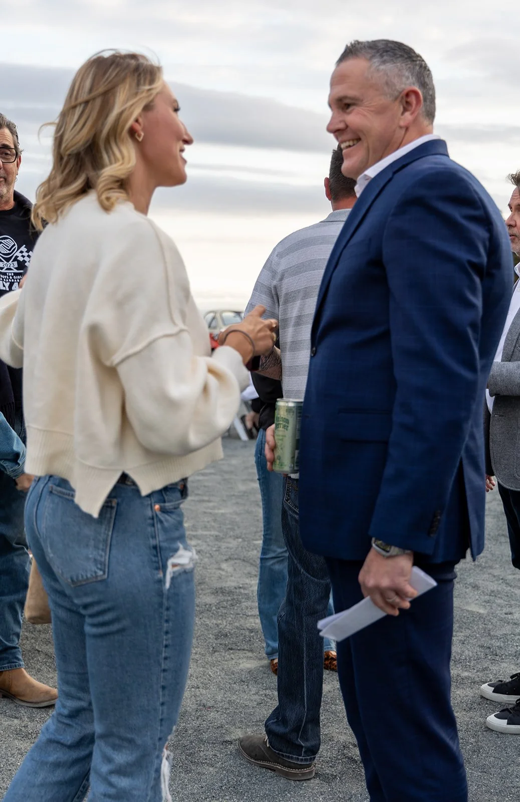 Two people, a woman and a man, are engaged in a conversation outdoors. The woman has blonde hair, is wearing a white sweater and ripped jeans, and is smiling. The man is dressed in a blue suit with a white shirt and is holding a soda can and some pap