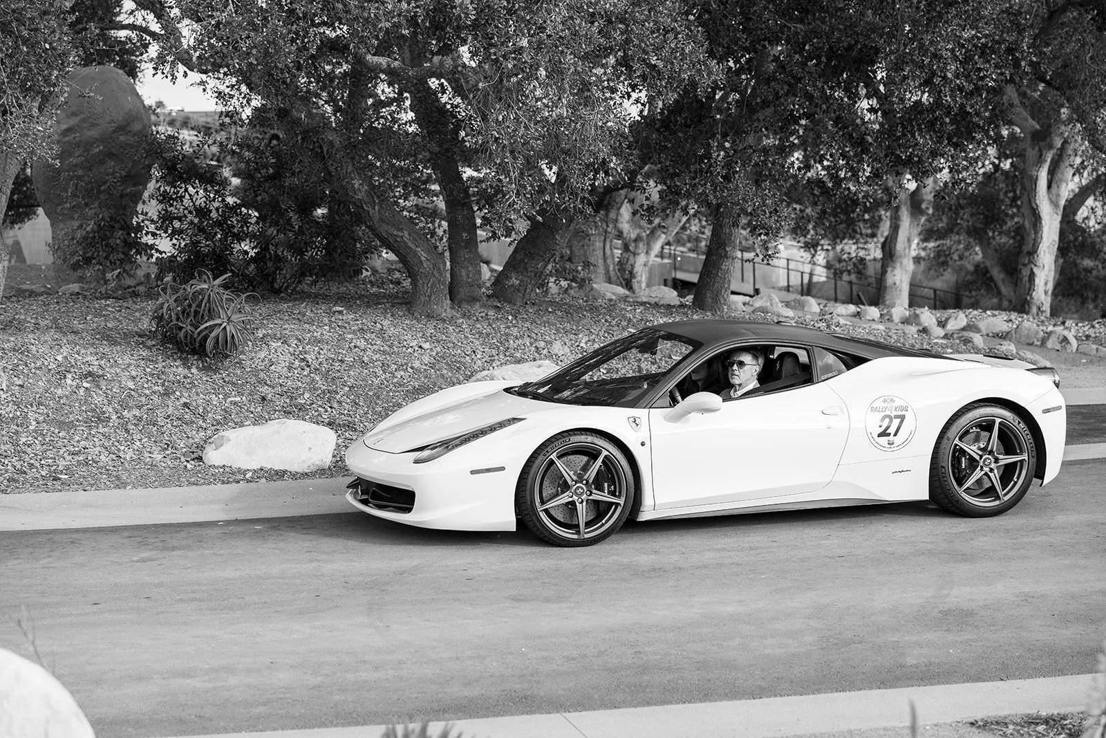 A white Ferrari sports car with the number 27 decal parked on a street, with a person inside wearing sunglasses, trees, rocks, and a sidewalk in the background.