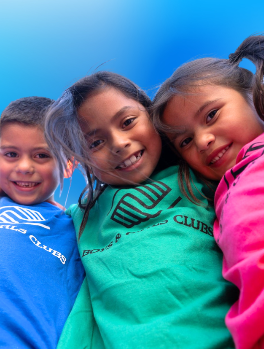 Three children smiling and looking down at the camera against a blue sky background.
