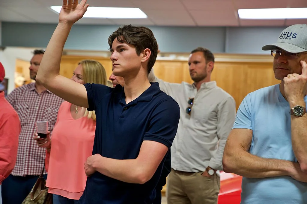 A young man in a navy blue polo raises his hand at a gathering, surrounded by other people in casual clothing, in an indoor setting.