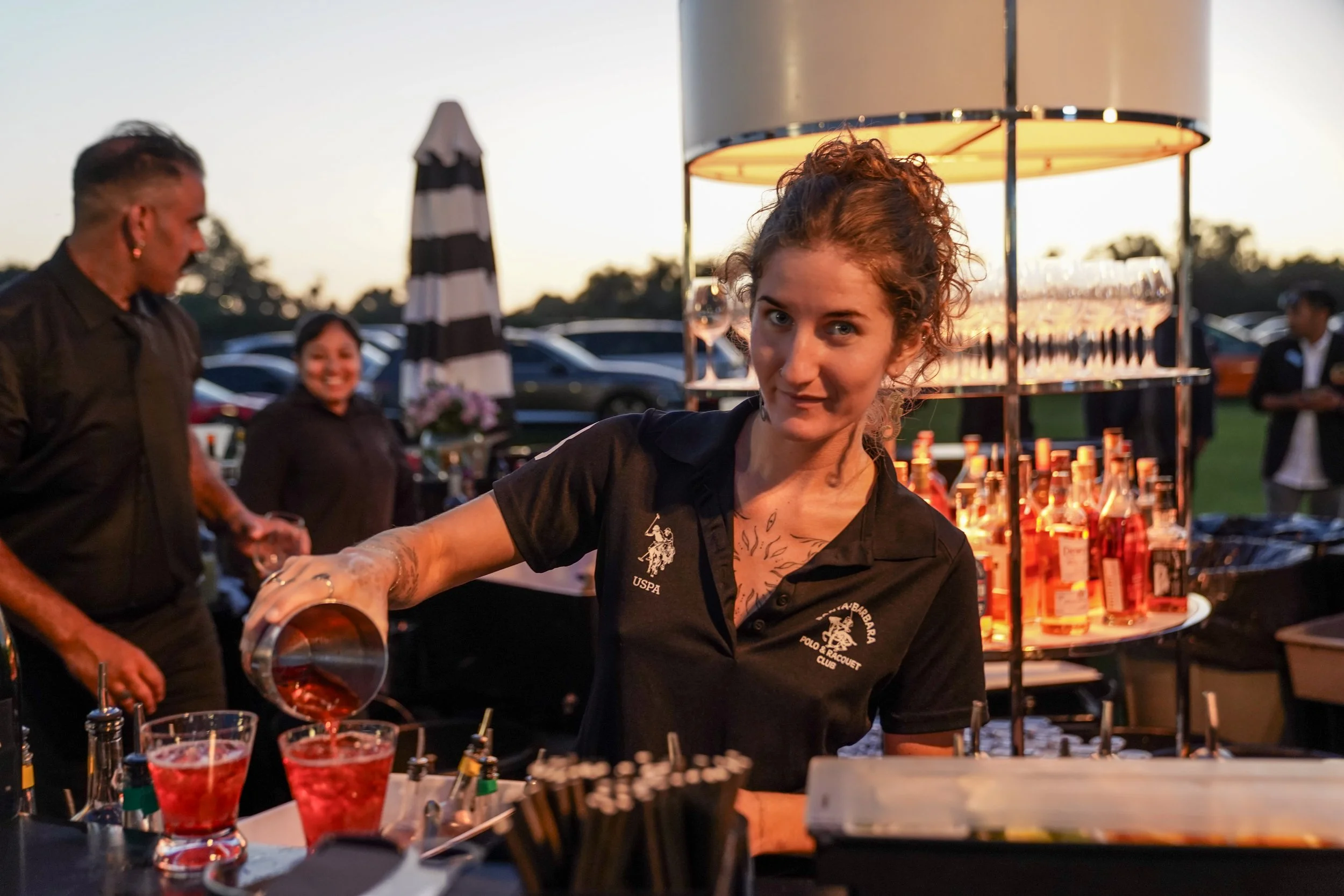A young woman pouring red cocktail into glasses at an outdoor event during sunset, with other people and parked cars in the background.