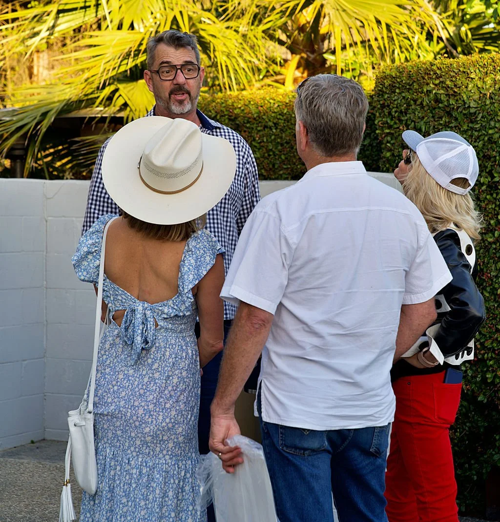 Four people engage in conversation outdoors, two men and two women, with one man wearing glasses and a checked shirt, a woman wearing a wide-brimmed hat and blue dress, a man wearing a white shirt, and a woman with blonde hair, a white cap, and red p