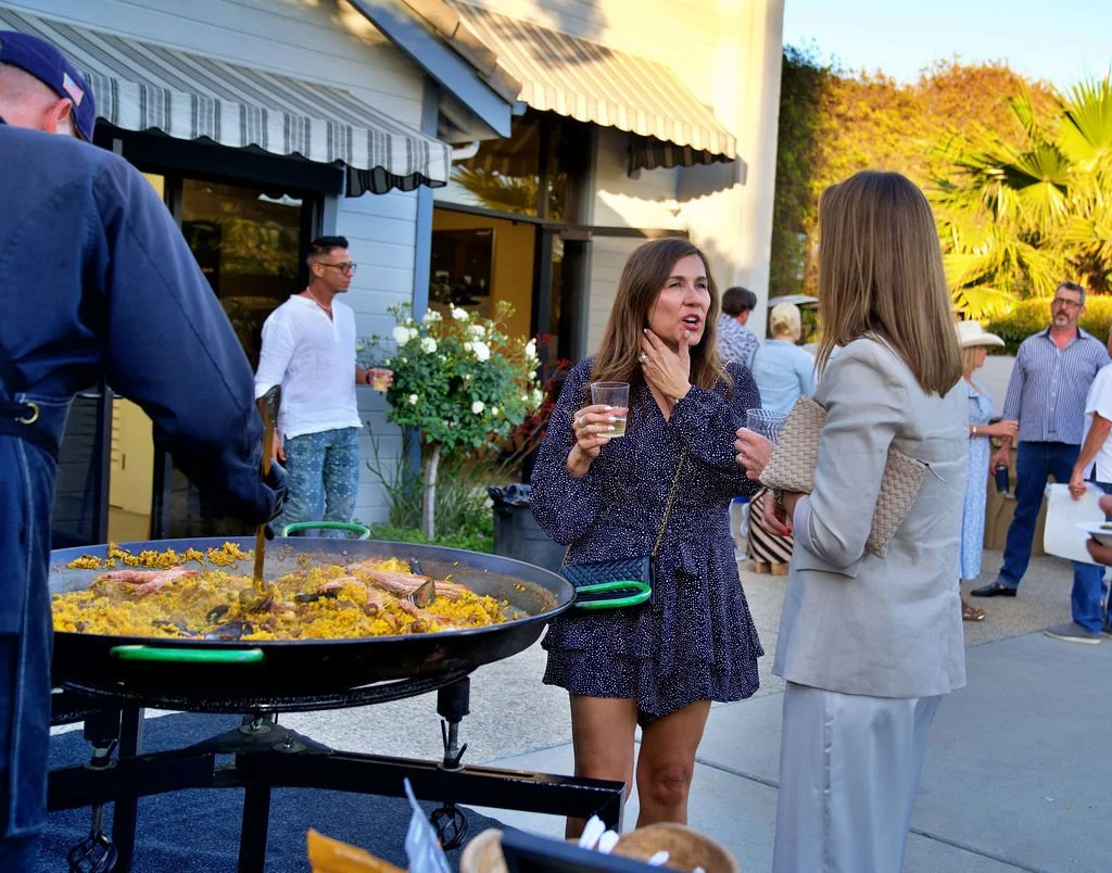 Two women enjoying conversation at an outdoor gathering, with a large paella dish in the foreground and other guests in the background.