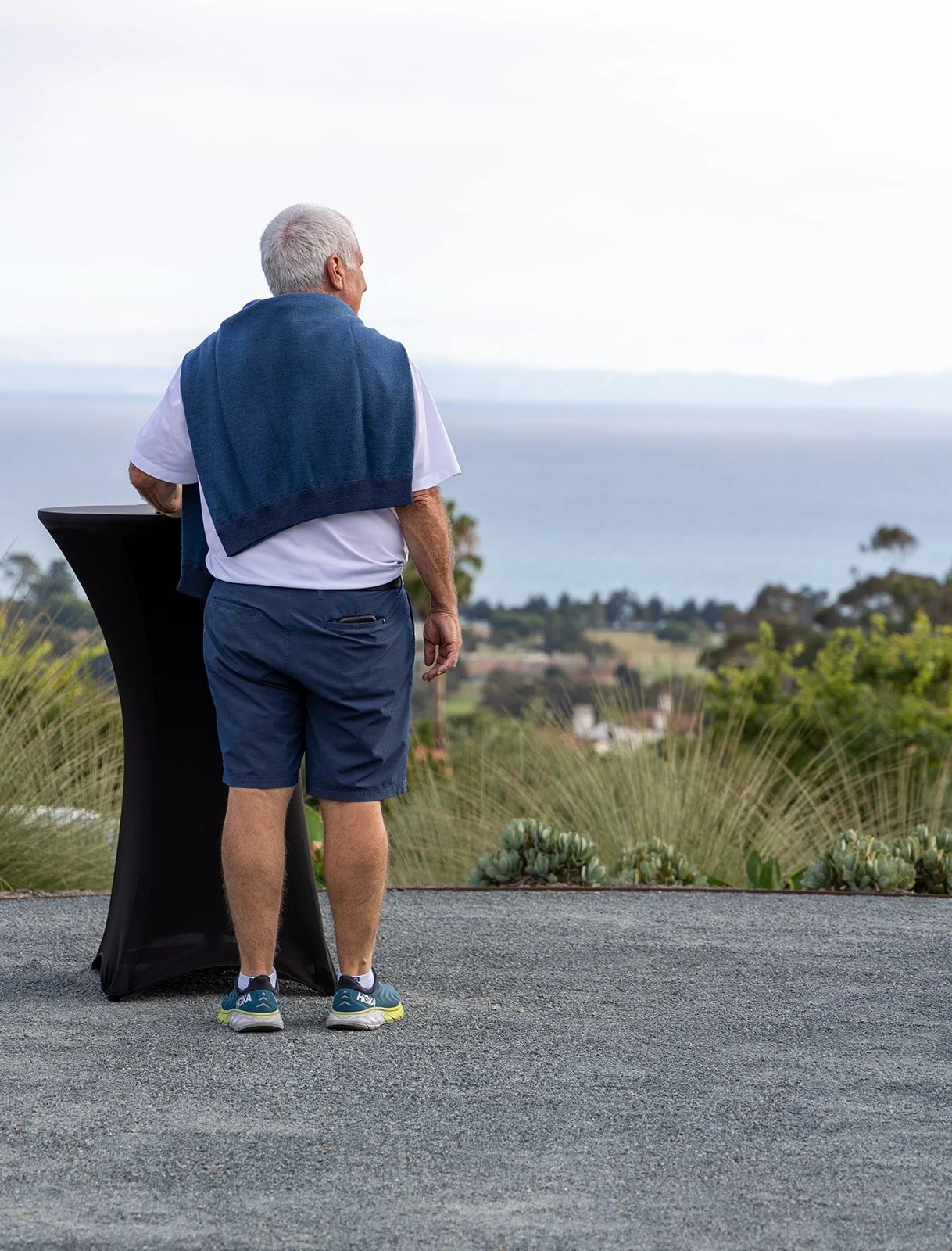 Older man with gray hair standing outdoors near a black standing table, overlooking a scenic view of the ocean and greenery, with a blue towel draped over his shoulders.