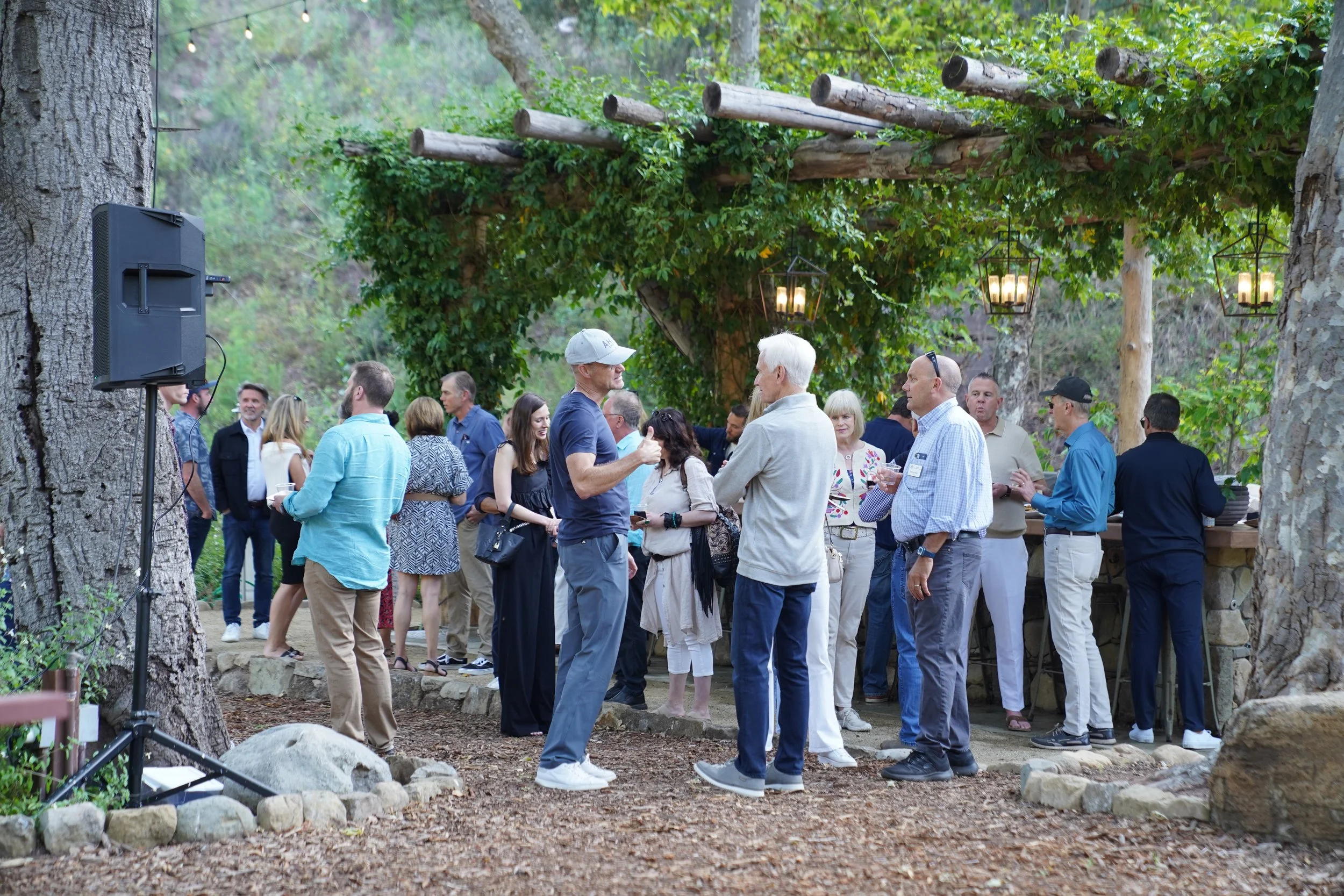 A group of people gathering outdoors in a wooded area, socializing at a rustic-style event with hanging lanterns and natural decor.