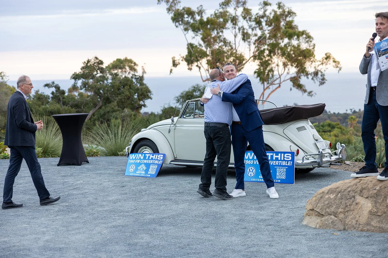 Two men in suits hugging in front of a vintage convertible car at an outdoor event, with two signs promoting a giveaway for a 1960 VW convertible, and other people socializing nearby against a scenic background of trees and a distant view of water or
