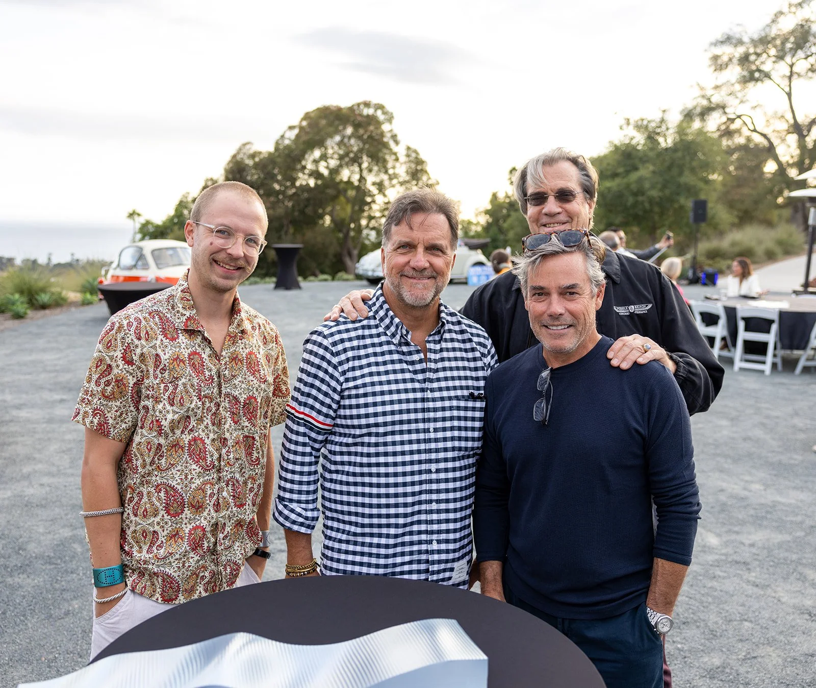 Four men standing outdoors at a gathering, smiling at the camera, with tables and trees in the background.