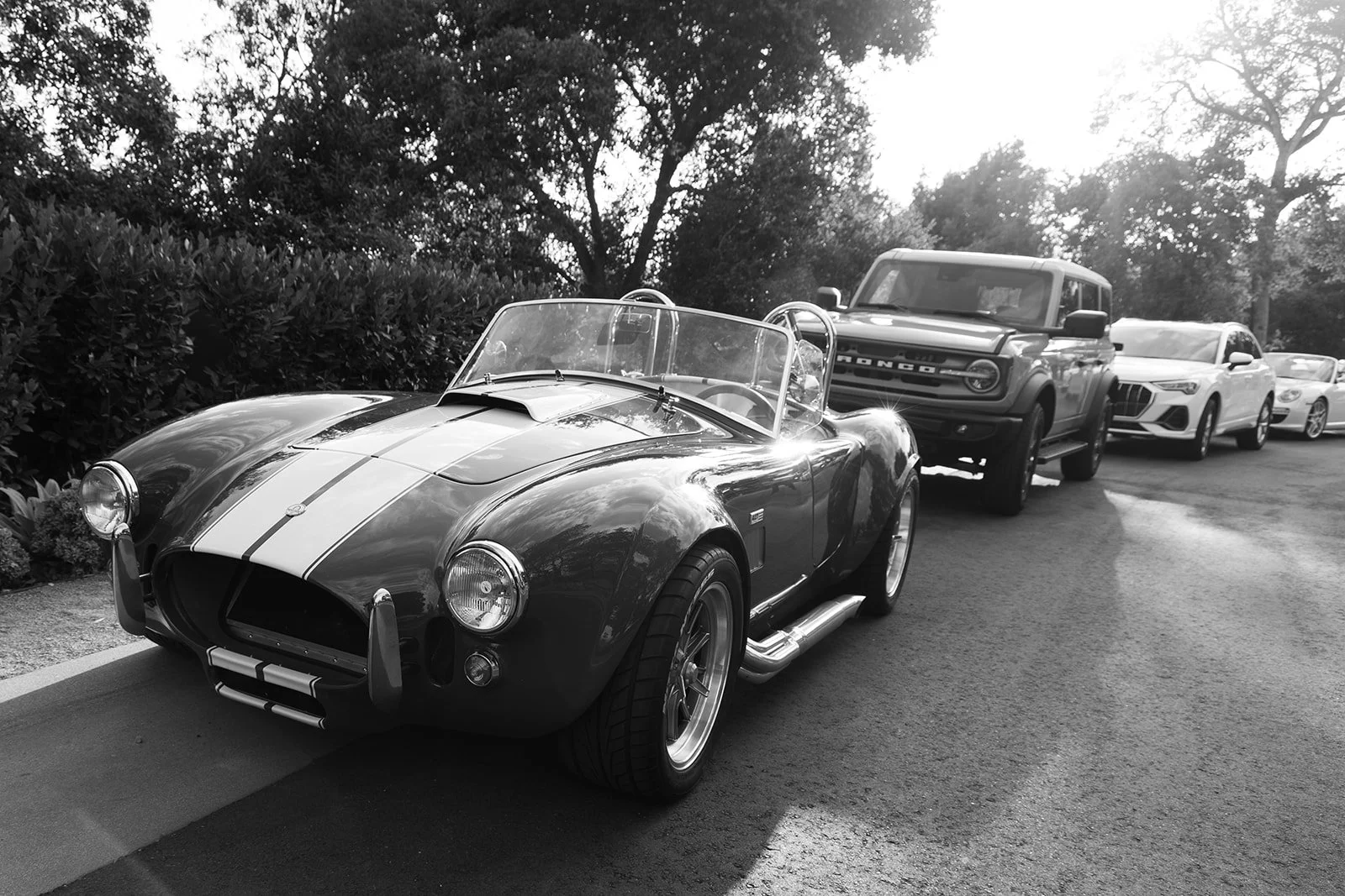 A line of four vehicles parked along a street, featuring a vintage sports car in the front and modern SUVs and cars behind it, with trees and bushes in the background.