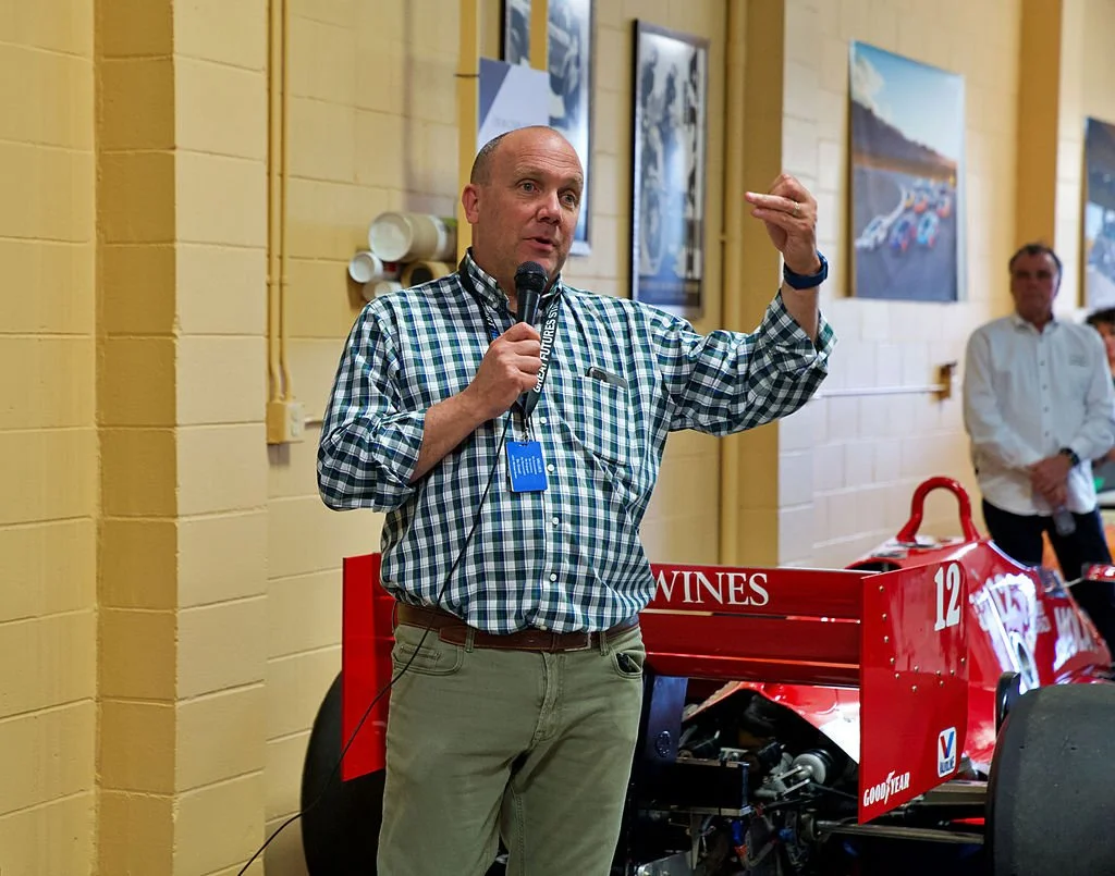 A man in a checkered shirt is speaking into a microphone, standing next to a red racing car in an indoor setting with framed pictures on the wall and other people in the background.