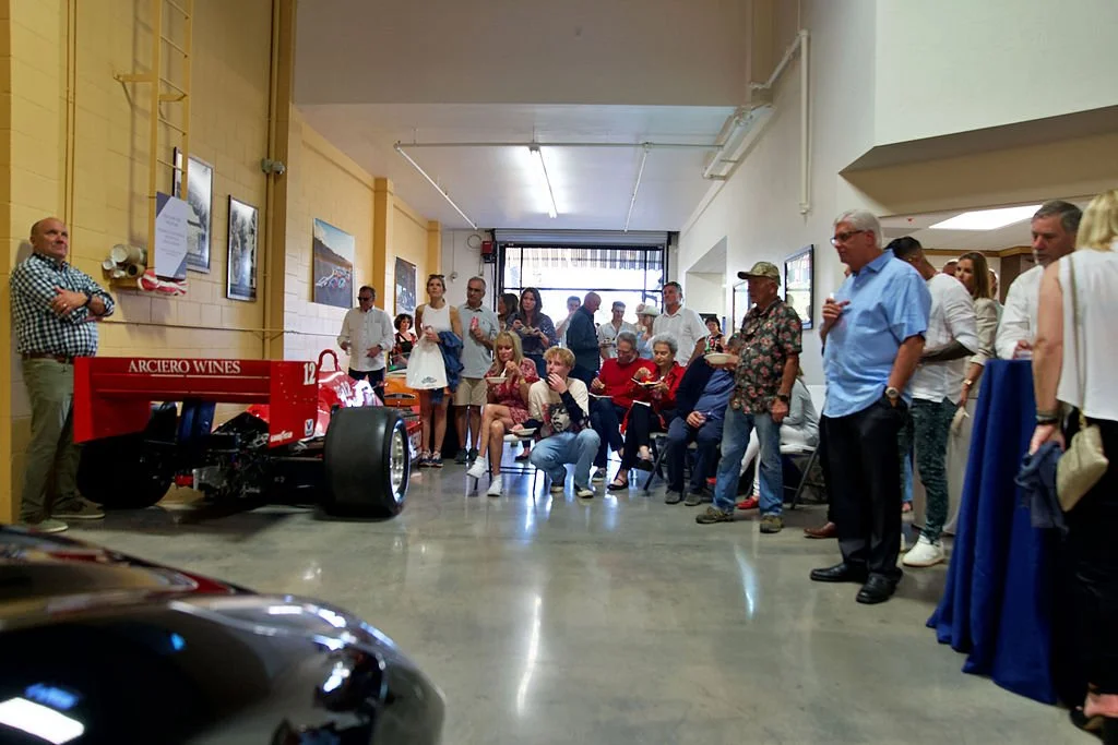 Group of people gathered indoors, standing and sitting, watching a red race car with 'ARCLOGO WINES' on the side, in what appears to be an exhibition or event space.