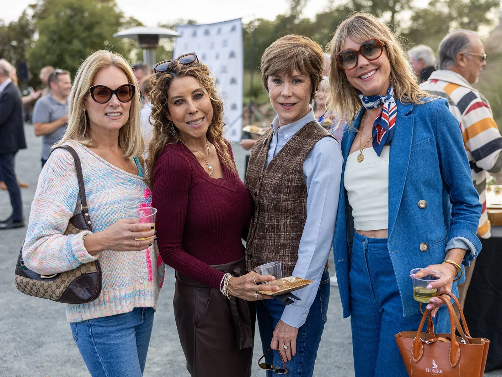Four women standing outdoors at a social event, smiling at the camera, with other people in the background.