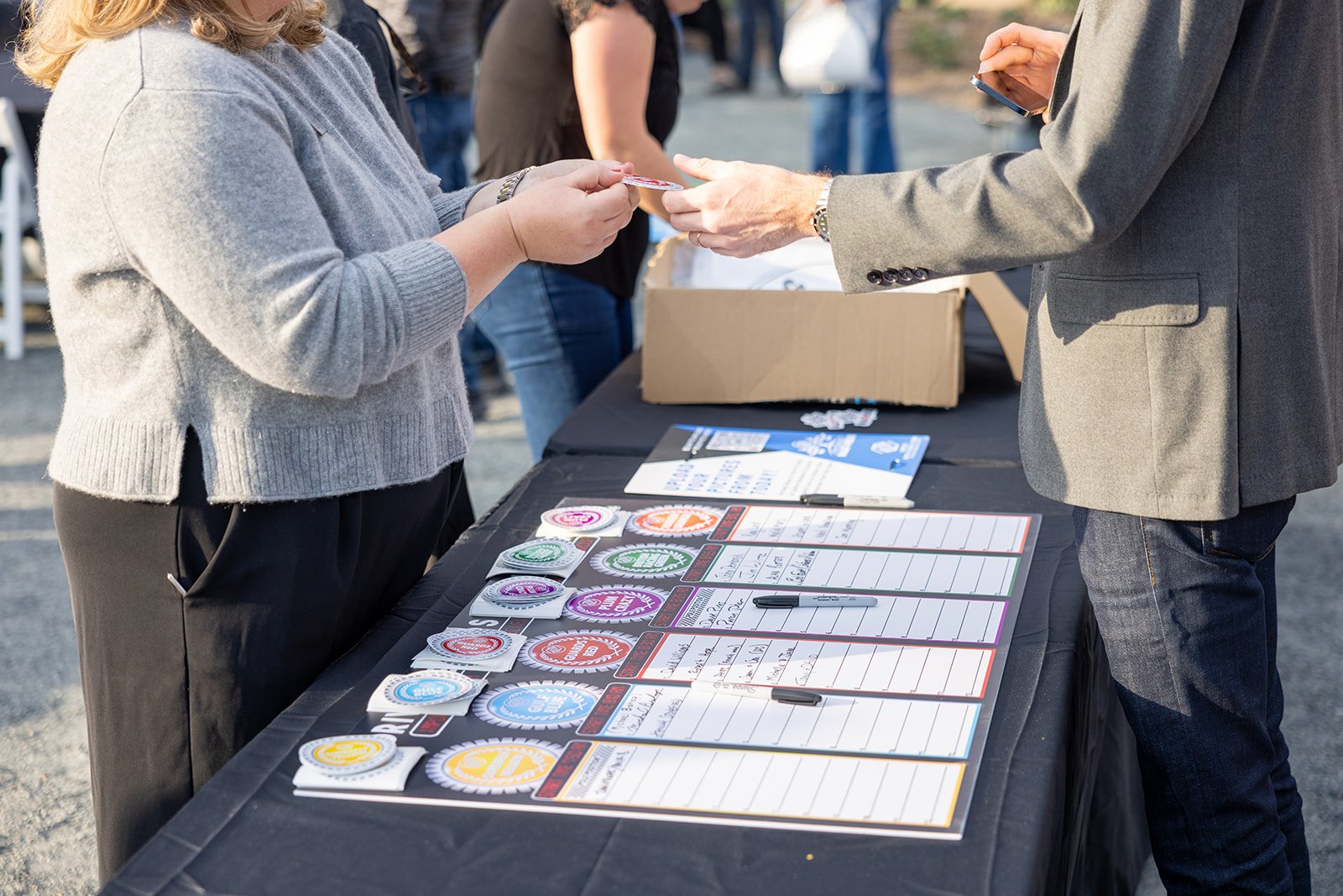 Two people exchanging a paper at a table covered with various colorful discount or promotional badges and clipboards with handwritten notes, outdoors with other people and a cardboard box in the background.