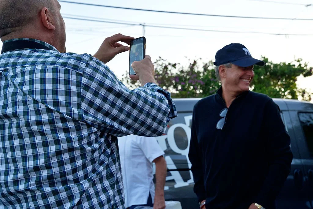 A man in a plaid shirt taking a photo of a woman in a black jacket, who is smiling and wearing a navy cap, outdoor with a gray vehicle and trees in the background.