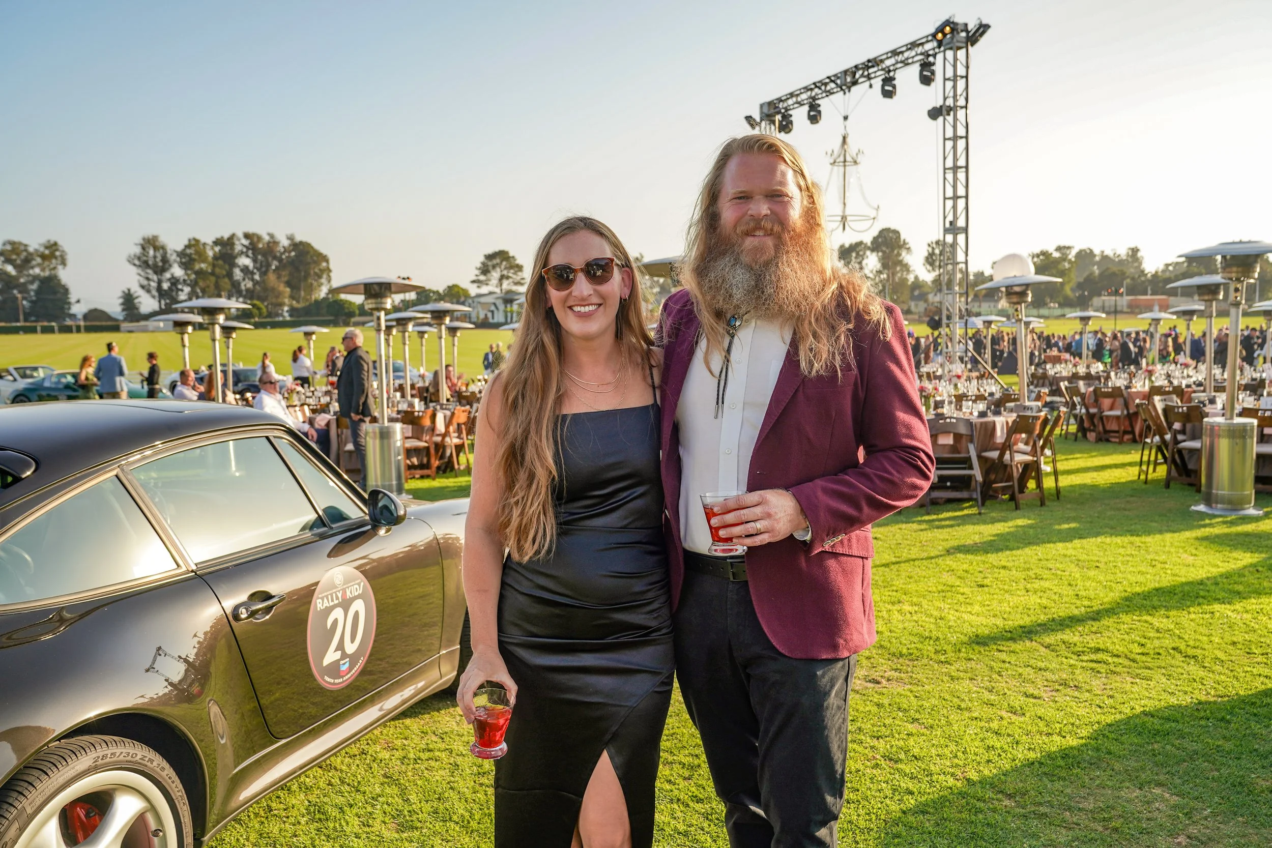 A man and woman standing together at an outdoor event, holding drinks, with a vintage car and large gathering of people in the background.