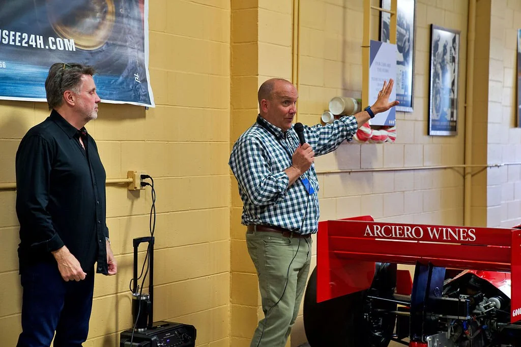 Two men standing in front of a yellow wall, one speaking into a microphone and gesturing towards framed pictures, with a red wine press labeled 'Arciero Wines' nearby.