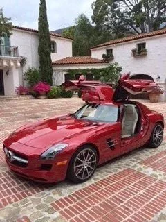 Red sports car with gullwing doors open, parked on a brick driveway in front of white buildings with tiled roofs, surrounded by greenery and pink flowers.