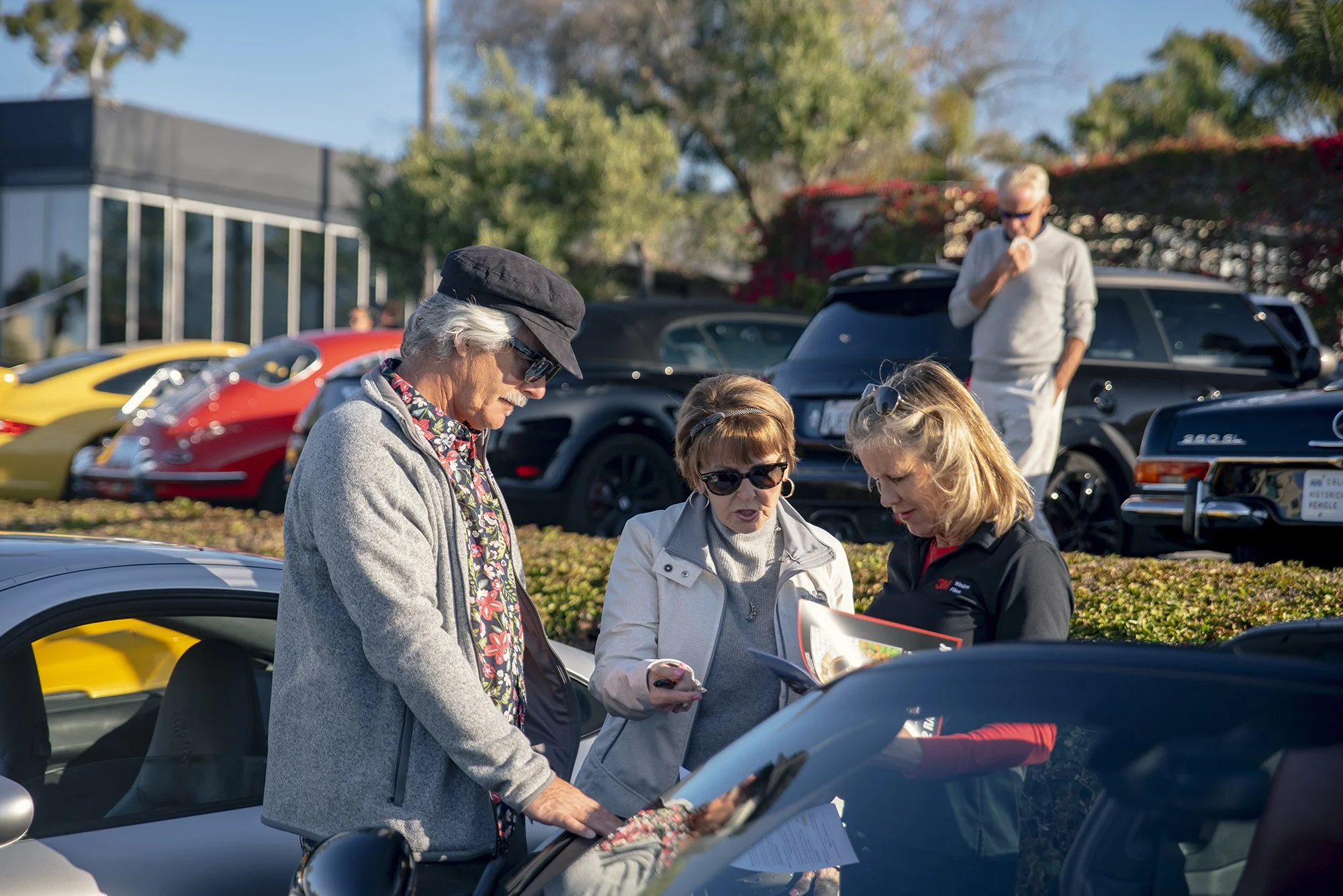 Three women are gathered around a silver sports car, looking at a brochure or book, with classic and luxury cars parked behind them on a sunny day.