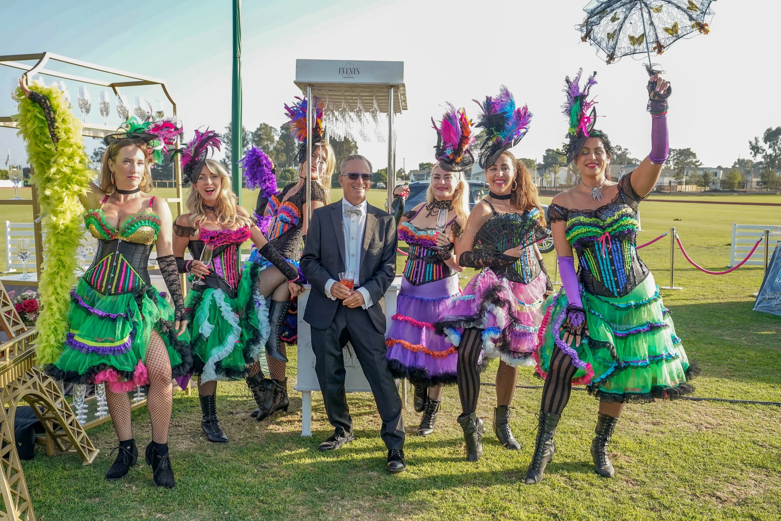 A group of women in colorful, feathered carnival costumes and a man in a tuxedo posing outdoors at an event.
