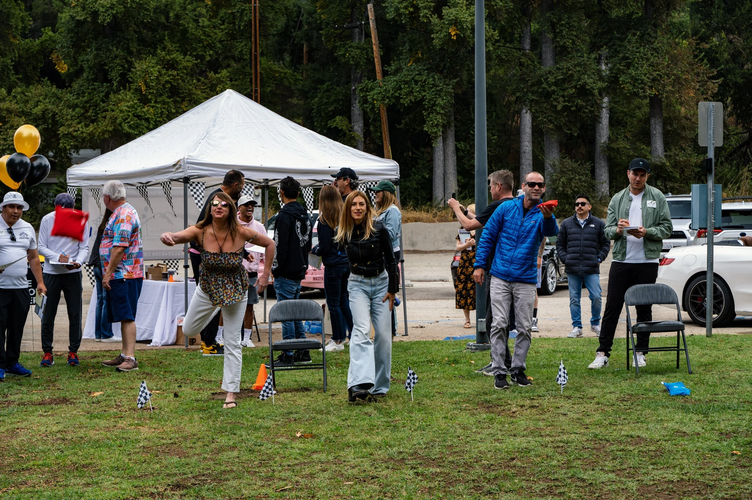 People gather outdoors at a event with checkered flags on the grass, some participating in an activity, others watching near a white tent and parked cars, surrounded by trees.