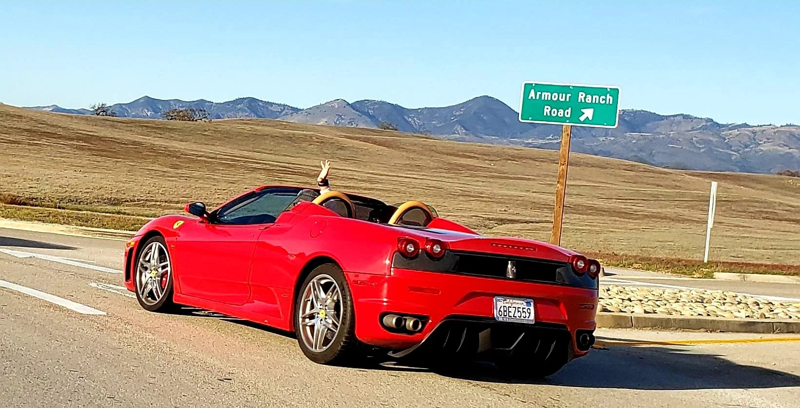 A red Ferrari convertible car parked on the side of a road with a driver waving from inside. There is a sign that reads 'Armour Ranch Road' and a mountain range in the background.