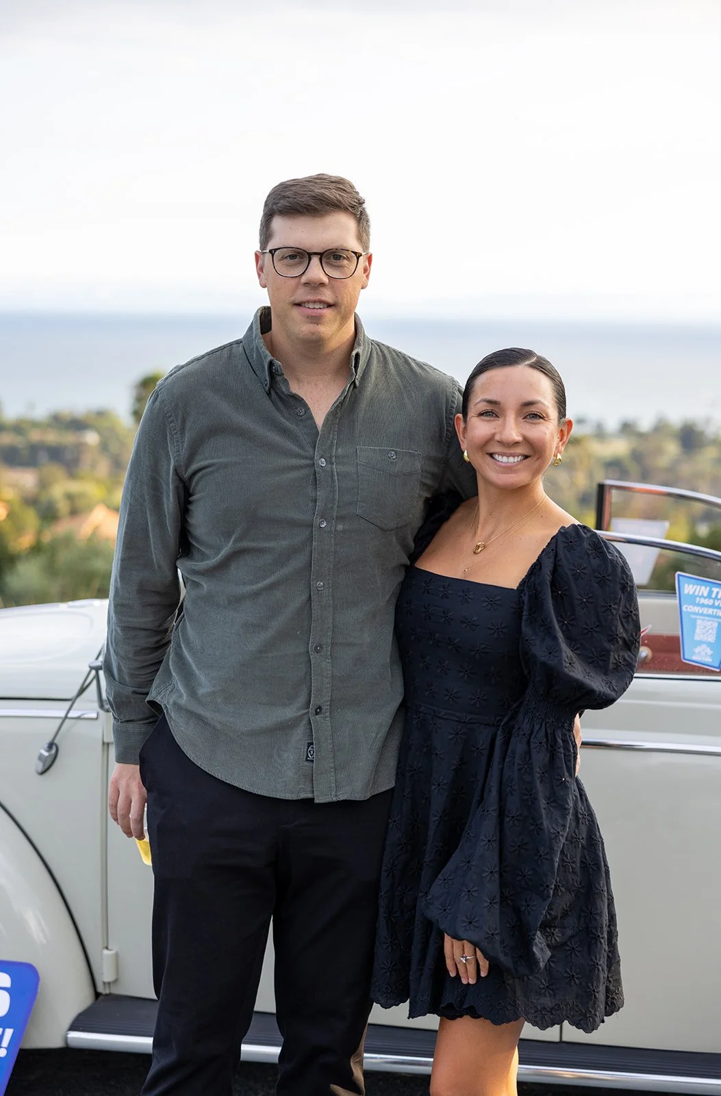 A man and woman standing side by side outdoors, smiling, with a scenic background of trees and a body of water, next to a vintage car.