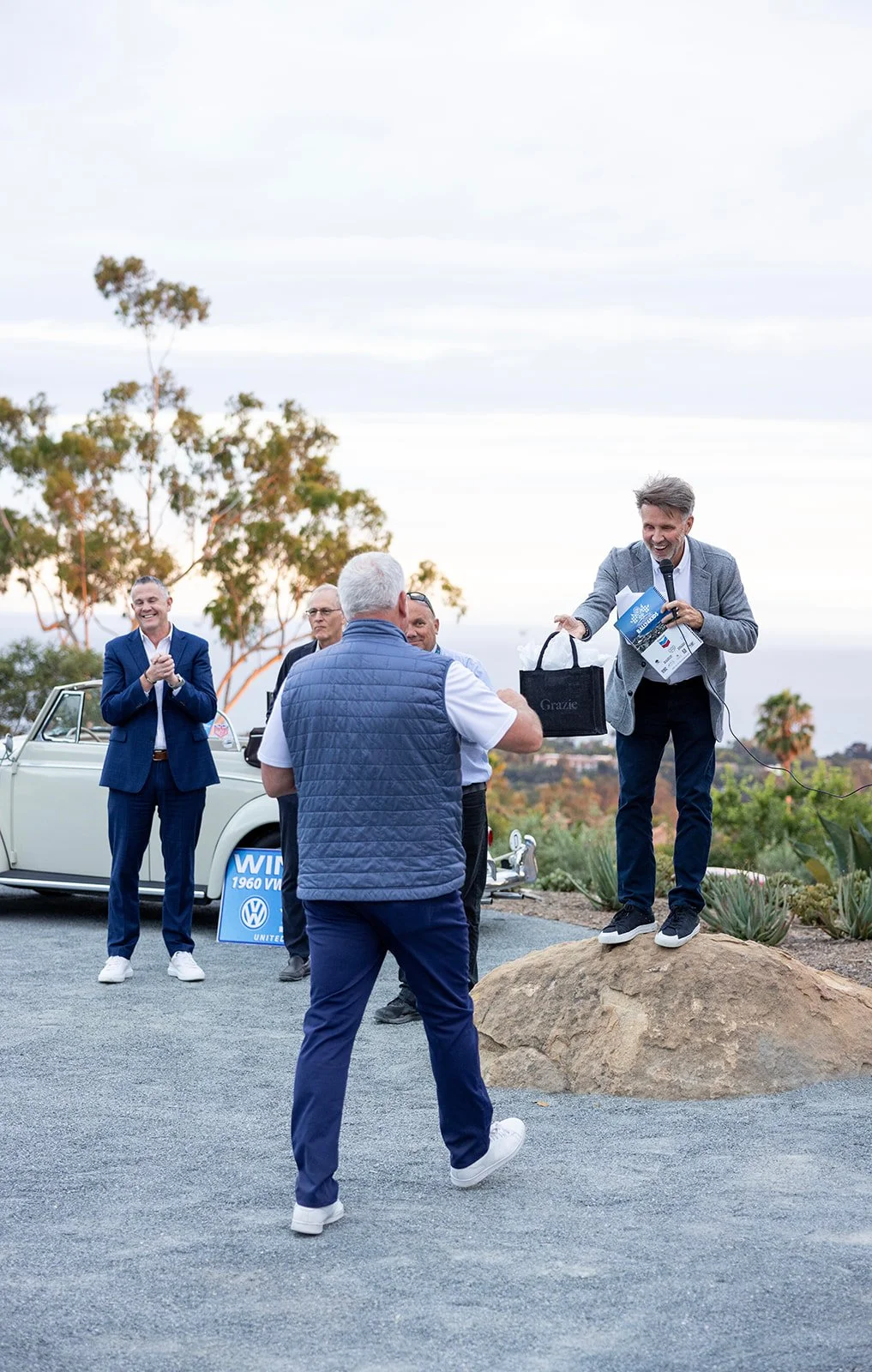 A man on a rock is holding a gift bag, smiling, and speaking into a microphone at an outdoor event with several other men clapping and smiling in the background. There is a vintage white car and a sign with the Volkswagen logo behind them.