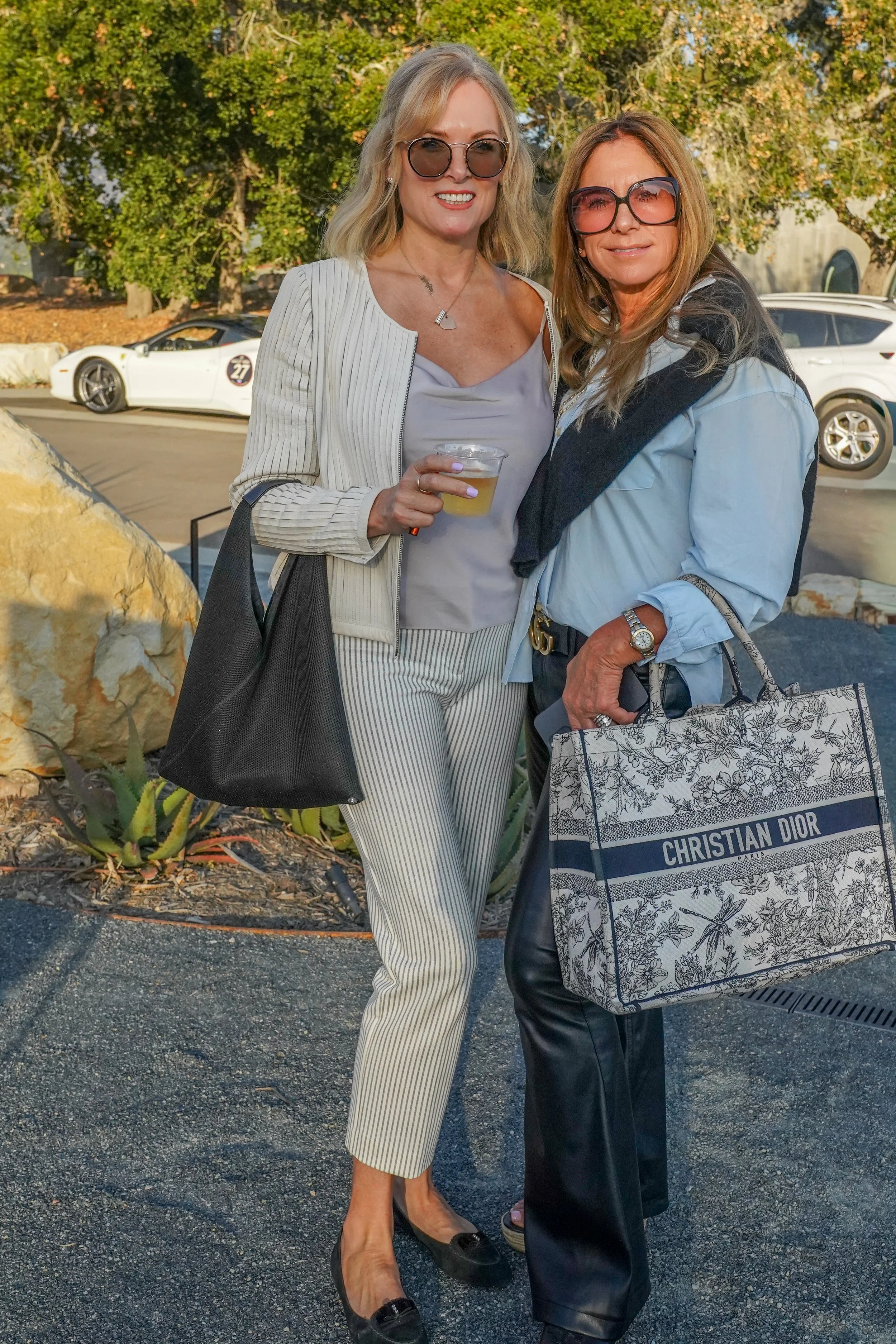 Two women standing outdoors, smiling, wearing sunglasses, and holding shopping bags and a drink, in a parking lot with trees and cars in the background.