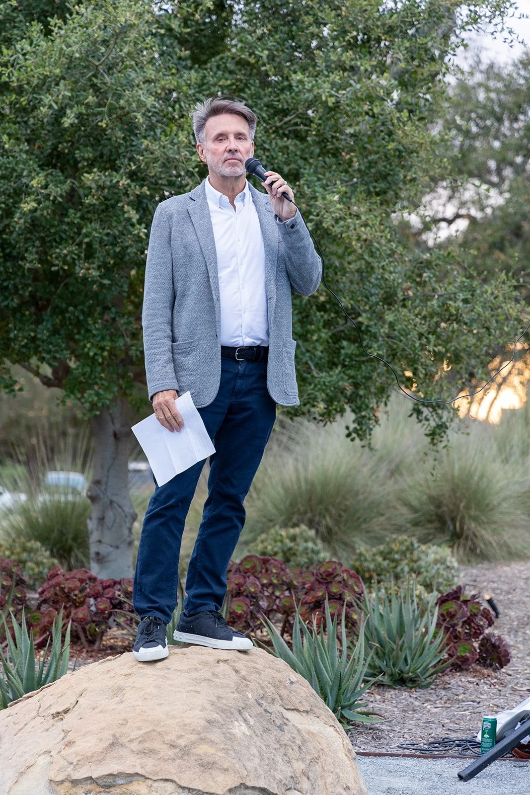 A man in a gray blazer, white shirt, and blue jeans standing on a large rock outdoors, holding a microphone in one hand and some papers in the other, with greenery in the background.